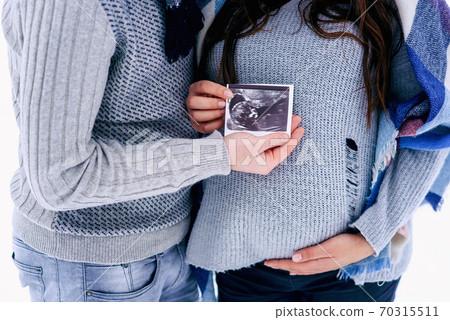 Close up man and his pregnant woman on warm clothes holding ultrasound pregnancy picture in their hands while they stand on snowy winter park. Close up man and his pregnant woman on warm clothes holding ultrasound pregnancy picture in their hands while they stand on snowy winter park. 70315511