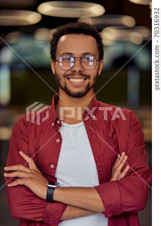 Vertical shot of a young happy mixed race businessman or office worker in casual wear keeping arms crossed, looking at camera and smiling while posing in the office 70316932