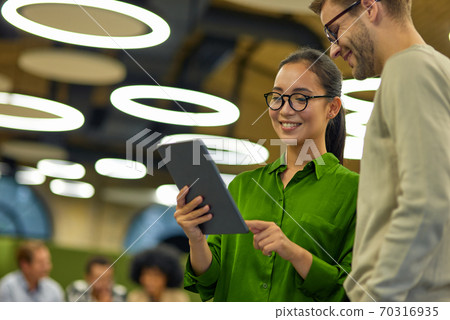 Young cheerful asian woman showing something on digital tablet to her male colleague while standing together in the coworking space or office Young cheerful asian woman showing something on digital tablet to her male colleague while standing together in the coworking space or office 70316935