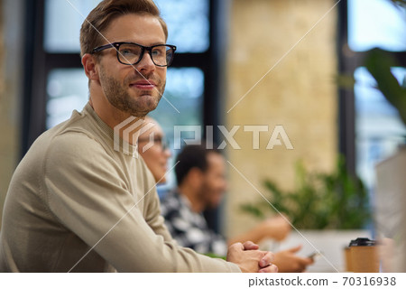 Young handsome man, male office worker sitting at desk in the board room and looking at camera while having a meeting with colleagues Young handsome man, male office worker sitting at desk in the board room and looking at camera while having a meeting with colleagues 70316938
