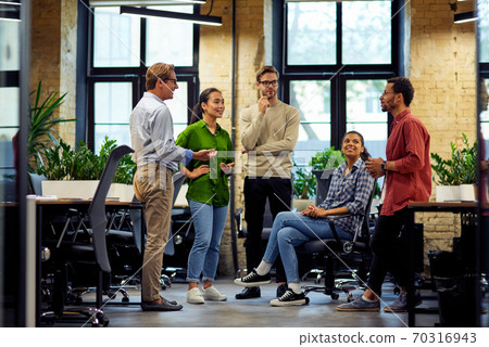 Brainstorming meeting. Group of young diverse multi ethnic business people standing together in modern office, sharing ideas and discussing work, full length Brainstorming meeting. Group of young diverse multi ethnic business people standing together in modern office, sharing ideas and discussing work, full length 70316943