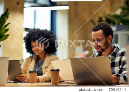 Using modern technologies. Two young multiracial business people sitting at the desk, working together in coworking space Using modern technologies. Two young multiracial business people sitting at the desk, working together in coworking space 70316944