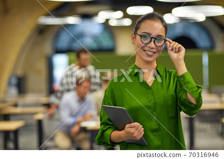Portrait of young happy asian woman adjusting her eyeglasses, holding digital tablet and smiling at camera while standing in the office with colleagues on the background Portrait of young happy asian woman adjusting her eyeglasses, holding digital tablet and smiling at camera while standing in the office with colleagues on the background 70316946