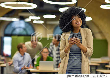 Young happy mixed race woman holding smartphone, looking aside and smiling while standing in the modern coworking space with her colleagues working on the background Young happy mixed race woman holding smartphone, looking aside and smiling while standing in the modern coworking space with her colleagues working on the background 70316947