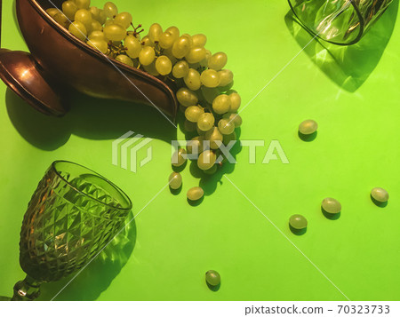 A bunch of white grapes and transparent green wine glasses on a green contrasting background and several grapes 70323733