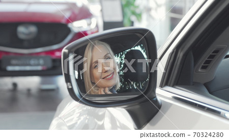 Beautiful happy woman smiling to the camera through side mirror of her car Beautiful happy woman smiling to the camera through side mirror of her car 70324280