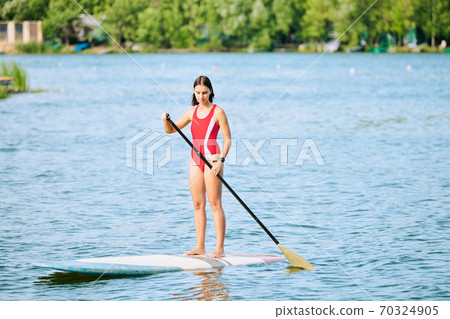 Young fit female surfboarder in swimsuit standing on surfboard while floating Young fit female surfboarder in swimsuit standing on surfboard while floating 70324905