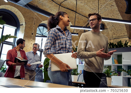 Two young diverse business people, male and female coworkers talking about something while walking in the modern office Two young diverse business people, male and female coworkers talking about something while walking in the modern office 70325630