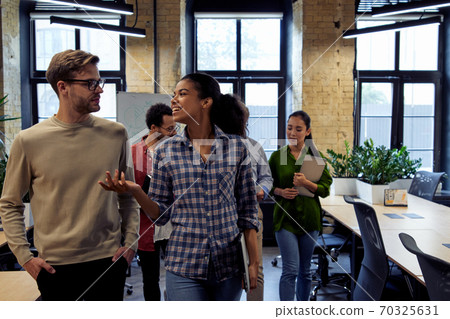 After team meeting. Two young diverse business people, male and female coworkers talking about something while walking in the modern office After team meeting. Two young diverse business people, male and female coworkers talking about something while walking in the modern office 70325631