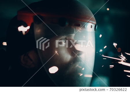 Man in transparent protective mask with flying sparks in darkness. Man in transparent protective mask with flying sparks in darkness. 70326622