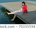 young beautiful woman sits on a bridge by the lake, legs down, sits in a red jacket 70335594