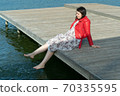 young beautiful woman sits on a bridge by the lake, legs down, sits in a red jacket 70335595