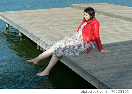 young beautiful woman sits on a bridge by the lake, legs down, sits in a red jacket young beautiful woman sits on a bridge by the lake, legs down, sits in a red jacket 70335595