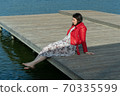 young beautiful woman sits on a bridge by the lake, legs down, sits in a red jacket 70335599