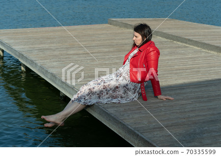 young beautiful woman sits on a bridge by the lake, legs down, sits in a red jacket 70335599