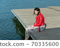 young beautiful woman sits on a bridge by the lake, legs down, sits in a red jacket 70335600