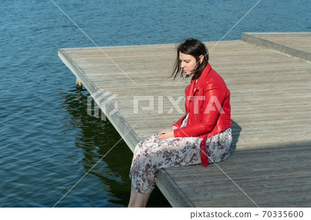young beautiful woman sits on a bridge by the lake, legs down, sits in a red jacket 70335600