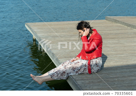 young beautiful woman sits on a bridge by the lake, legs down, sits in a red jacket young beautiful woman sits on a bridge by the lake, legs down, sits in a red jacket 70335601