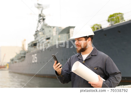 Marine engineer talking by VHF walkie talkie, holding papers near vessel in background. 70338792