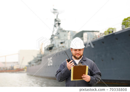 Marine assistant engineer talking by walkie talkie radio and standing near vessel with documents in hands. 70338808