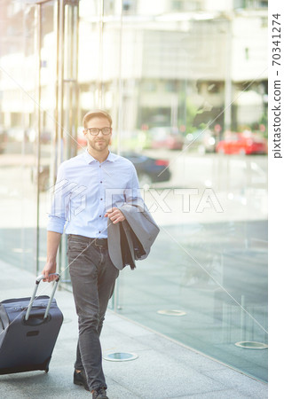 Going to airport terminal. Vertical shot of a young confident businessman walking on city streets and pulling his suitcase Going to airport terminal. Vertical shot of a young confident businessman walking on city streets and pulling his suitcase 70341274