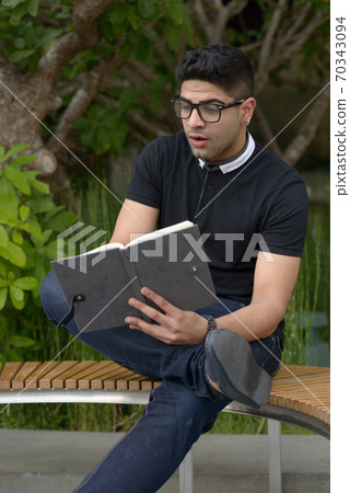 Young handsome Indian man with eyeglasses reading book and looking shocked at the park Young handsome Indian man with eyeglasses reading book and looking shocked at the park 70343094