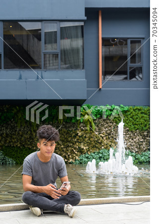 Young Asian man with phone sitting outside modern building in the city Young Asian man with phone sitting outside modern building in the city 70345094