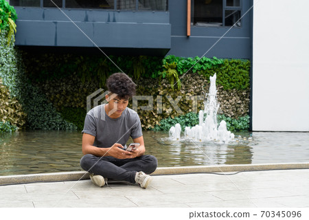 Young Asian man sitting while using phone outside modern building Young Asian man sitting while using phone outside modern building 70345096
