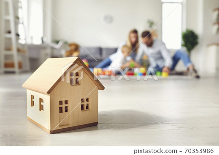 Little wooden house on floor of cozy room with happy family playing in background Little wooden house on floor of cozy room with happy family playing in background 70353986