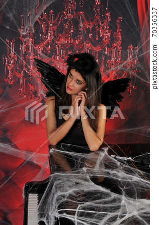 Beautiful young brunette woman in black angel costume with wings over spooky red background posing by piano with rose bouquet. Halloween party outfit 70356337