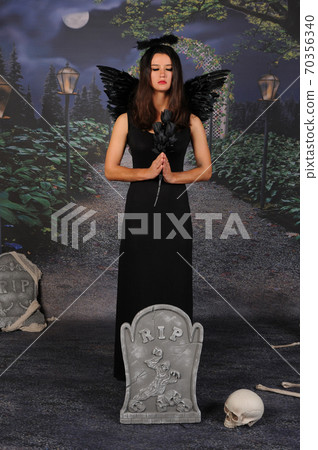 Beautiful young brunette woman in black angel costume with wings over dark spooky background in praying pose. Halloween party outfit 70356340