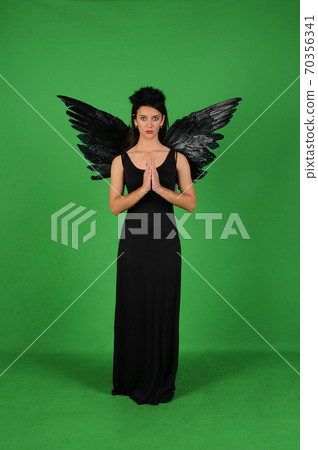 Beautiful young brunette woman in black angel costume with wings over green background in praying pose. Halloween party outfit 70356341