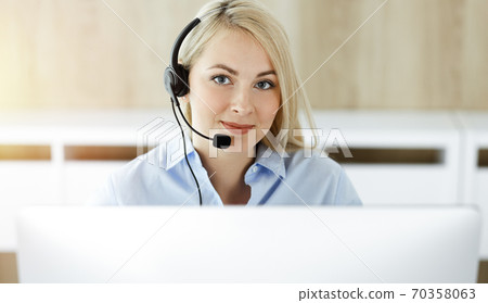 Blonde business woman sitting and communicated by headset in call center in sunny office. Concept of telesales business 70358063