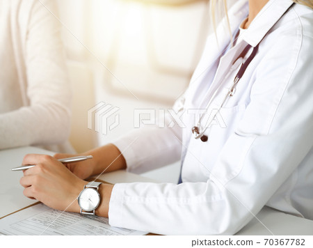 Unknown woman-doctor communicating with female patient in sunny clinic. Physician checks medical history record and exam results, close-up 70367782