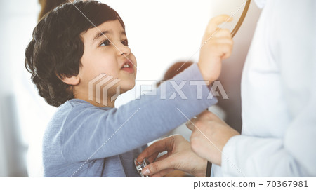 Doctor-woman examining a child patient by stethoscope in sunny clinik. Cute arab boy and his brother at physician appointment 70367981
