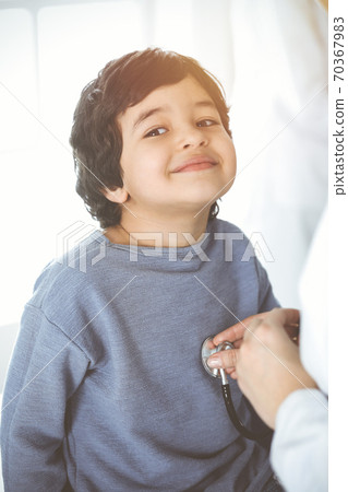 Doctor-woman examining a child patient by stethoscope in sunny clinik. Cute arab boy and his brother at physician appointment 70367983