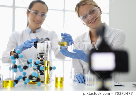 Two women in uniform and chemical goggles hold glass flask of yellow liquid in front of camera 70373477