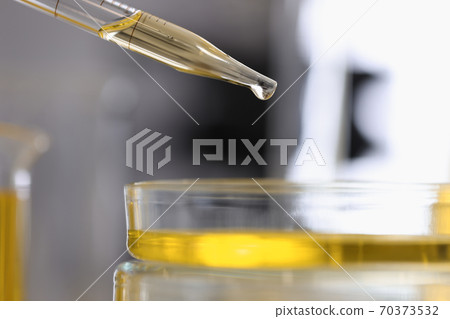 Dispenser filled with yellow chemical liquid pours drop into a glass dish closeup 70373532