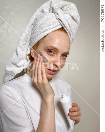 Beautiful woman after a shower in a towel on her head wants to apply a cosmetic mask on her face at home 70376971