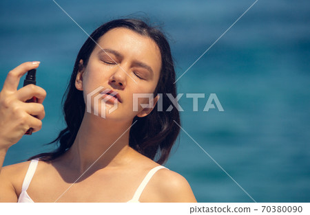 Young beautiful woman spraying thermal water or facial mist on her face at the beach. Summer skin care Young beautiful woman spraying thermal water or facial mist on her face at the beach. Summer skin care 70380090