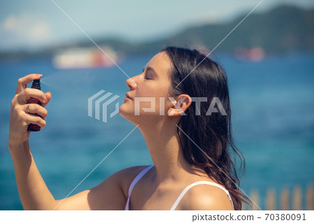 Young beautiful woman spraying thermal water or facial mist on her face at the beach. Summer skin care Young beautiful woman spraying thermal water or facial mist on her face at the beach. Summer skin care 70380091