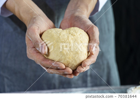 Close up hand of chef man wearing grey apron while holding homemade raw dough in heart shape. Close up hand of chef man wearing grey apron while holding homemade raw dough in heart shape. 70381644