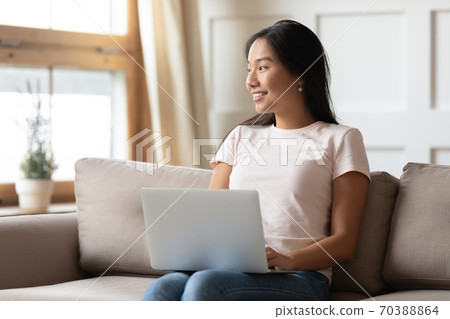 Smiling dreamy Asian young woman with laptop sitting on couch Smiling dreamy Asian young woman with laptop sitting on couch 70388864