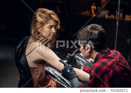 Modern experienced girl tattoo master makes a tattoo on the arm of a young worried girl with red hair. Modern experienced girl tattoo master makes a tattoo on the arm of a young worried girl with red hair. 70392671