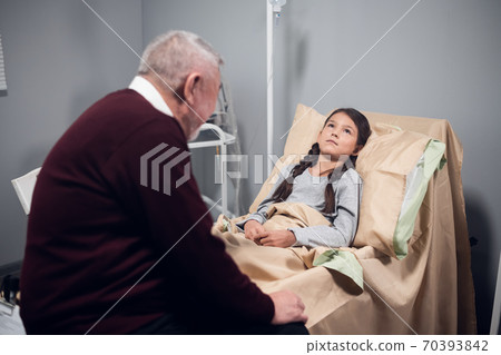 An old man is telling a kid a story in a hospital, while the kid is staying in bed and listening with an interest 70393842