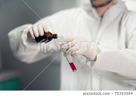 A male chemist holds a test tube of glass in his hand with some pink liquid and pours a new reagent into it A male chemist holds a test tube of glass in his hand with some pink liquid and pours a new reagent into it 70393884