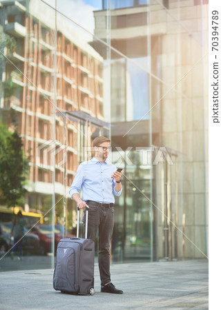 Vertical shot of elegant handsome businessman with suitcase using smartphone and looking away while standing outdoors, waiting for taxi 70394789