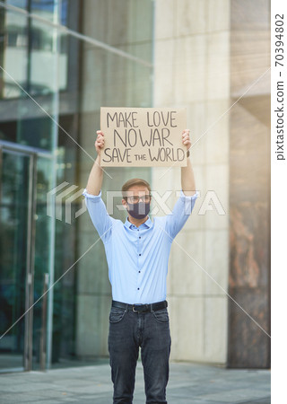 Save the world. Vertical of young male activist wearing black protective mask holding banner or poster and while protesting against the building outdoors Save the world. Vertical of young male activist wearing black protective mask holding banner or poster and while protesting against the building outdoors 70394802