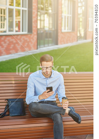 Writing business email. Vertical shot of a handsome businessman using smartphone and drinking coffee while sitting on the bench outdoors 70394810
