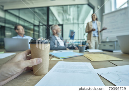 Close up shot of a male hand holding paper cup, man sitting at the business meeting in the modern office while young woman standing near flipchart and giving presentation Close up shot of a male hand holding paper cup, man sitting at the business meeting in the modern office while young woman standing near flipchart and giving presentation 70396729